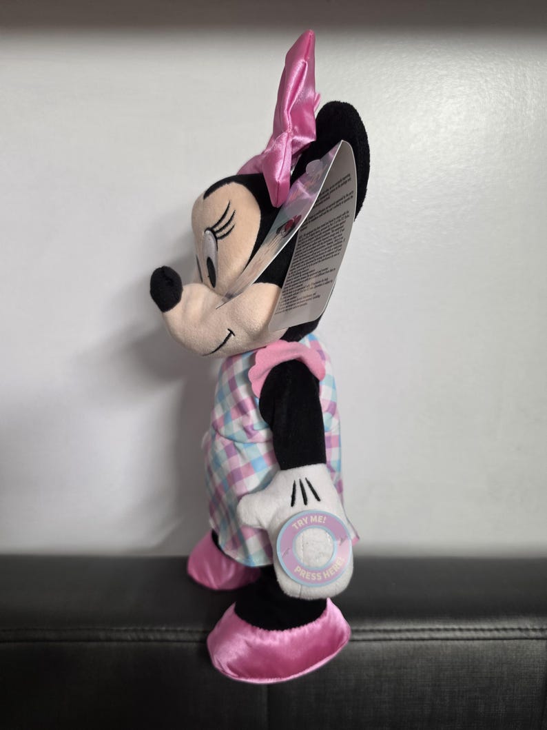 May include: A plush Minnie Mouse doll, featuring a black head, ears, and a pink bow. She wears a plaid dress in pastel colors and pink shoes. The doll has a white glove with a "TRY ME! PRESS HERE" tag.
