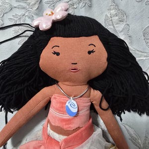 May include: A handmade doll with black yarn hair, a pink flower hair accessory, and a tan face. The doll wears a pink top and skirt, and a blue and white pendant necklace. The doll's arms are extended, and the background is a light blue floral pattern.