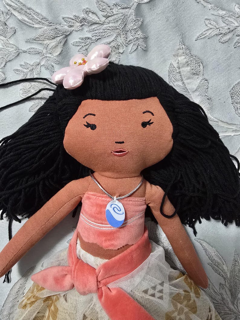 May include: A handmade doll with black yarn hair, a pink flower hair accessory, and a tan face. The doll wears a pink top and skirt, and a blue and white pendant necklace. The doll's arms are extended, and the background is a light blue floral pattern.