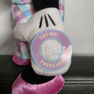 May include: A plush Minnie Mouse doll with a white glove holding a circular tag that says "TRY ME! PRESS HERE!". The doll is wearing a pink and blue plaid outfit and pink shoes. The doll's black shoe is visible.