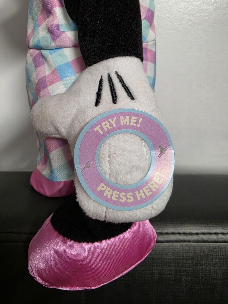 May include: A plush Minnie Mouse doll with a white glove holding a circular tag that says "TRY ME! PRESS HERE!". The doll is wearing a pink and blue plaid outfit and pink shoes. The doll's black shoe is visible.