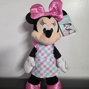 May include: A Minnie Mouse plush doll with a large pink bow, wearing a checkered apron and pink shoes. The doll has black ears and a tag that reads "Mickey and Friends".