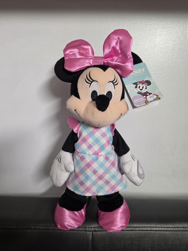 May include: A Minnie Mouse plush doll with a large pink bow, wearing a checkered apron and pink shoes. The doll has black ears and a tag that reads "Mickey and Friends".