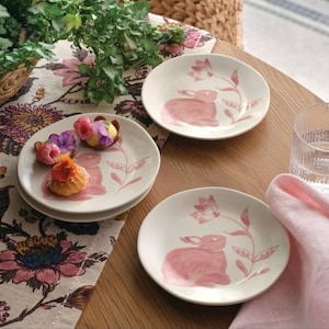 May include: Set of three white ceramic plates with a pink bunny and floral design. The plates are arranged on a wooden table with a floral patterned tablecloth and a pink napkin. The plates are decorated with a pink bunny and floral design.