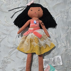 May include: A plush doll with black hair, wearing a pink and white top, a yellow skirt with white lace overlay, and a pink bow. The doll has a necklace with a blue pendant. A tag from "pill wfort" is attached.