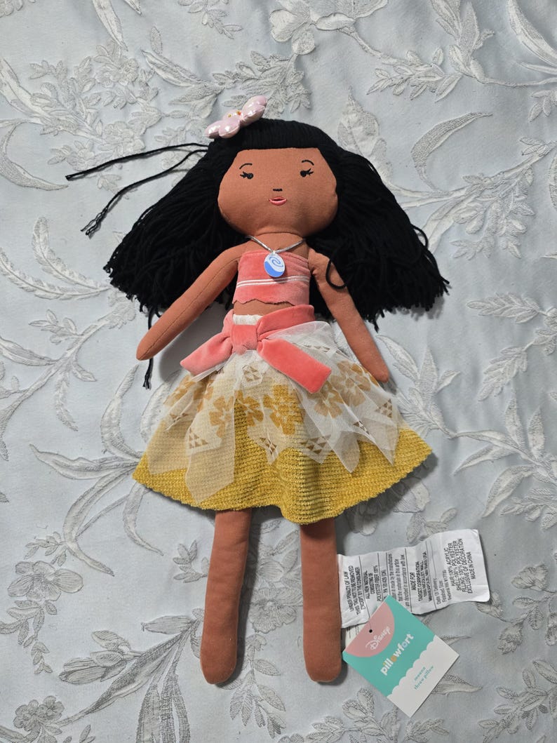 May include: A plush doll with black hair, wearing a pink and white top, a yellow skirt with white lace overlay, and a pink bow. The doll has a necklace with a blue pendant. A tag from "pill wfort" is attached.