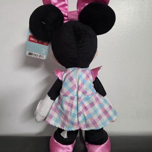 May include: A plush Minnie Mouse doll, viewed from the back, with black ears and a pink bow. She wears a plaid dress in shades of pink and blue, with matching pink shoes. A tag is attached to the doll.