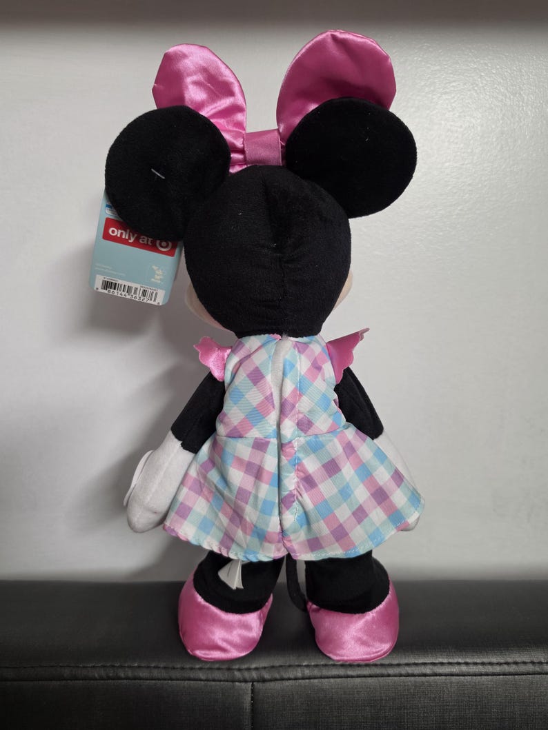 May include: A plush Minnie Mouse doll, viewed from the back, with black ears and a pink bow. She wears a plaid dress in shades of pink and blue, with matching pink shoes. A tag is attached to the doll.