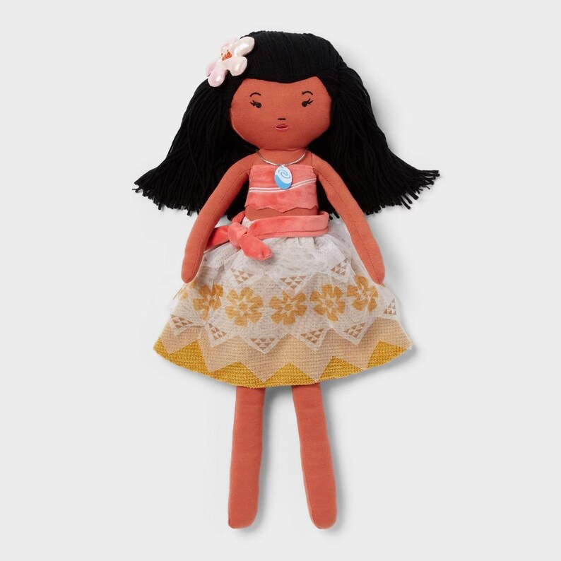 May include: A plush doll with black hair, a flower hair accessory, and a coral-colored skin tone. The doll wears a coral and white top, a white skirt with yellow floral patterns, and a blue pendant necklace. The doll's arms and legs are coral-colored.