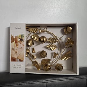 Op de afbeelding: Een goudkleurige metalen slinger met bloemen- en bladerontwerpen, weergegeven in een witte doos. De slinger heeft meerdere bloem- en bladvormen. De doos is gemarkeerd met "Garland" en "Threshold".