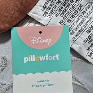 May include: A close-up of a teal and pink Disney Pillowfort tag for a Moana throw pillow. The tag has the Disney logo and the words "pillowfort" and "moana throw pillow". The tag is attached to a brown pillow.