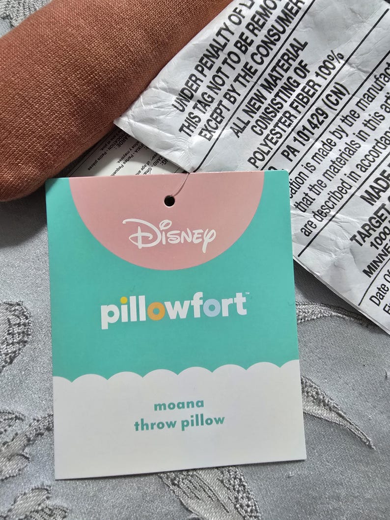 May include: A close-up of a teal and pink Disney Pillowfort tag for a Moana throw pillow. The tag has the Disney logo and the words "pillowfort" and "moana throw pillow". The tag is attached to a brown pillow.