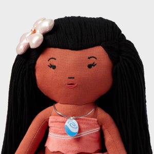 May include: A plush doll with dark hair and a pink dress. The doll has a flower hair accessory and a necklace with a blue and white pendant. The doll's skin is a warm, reddish-brown color.
