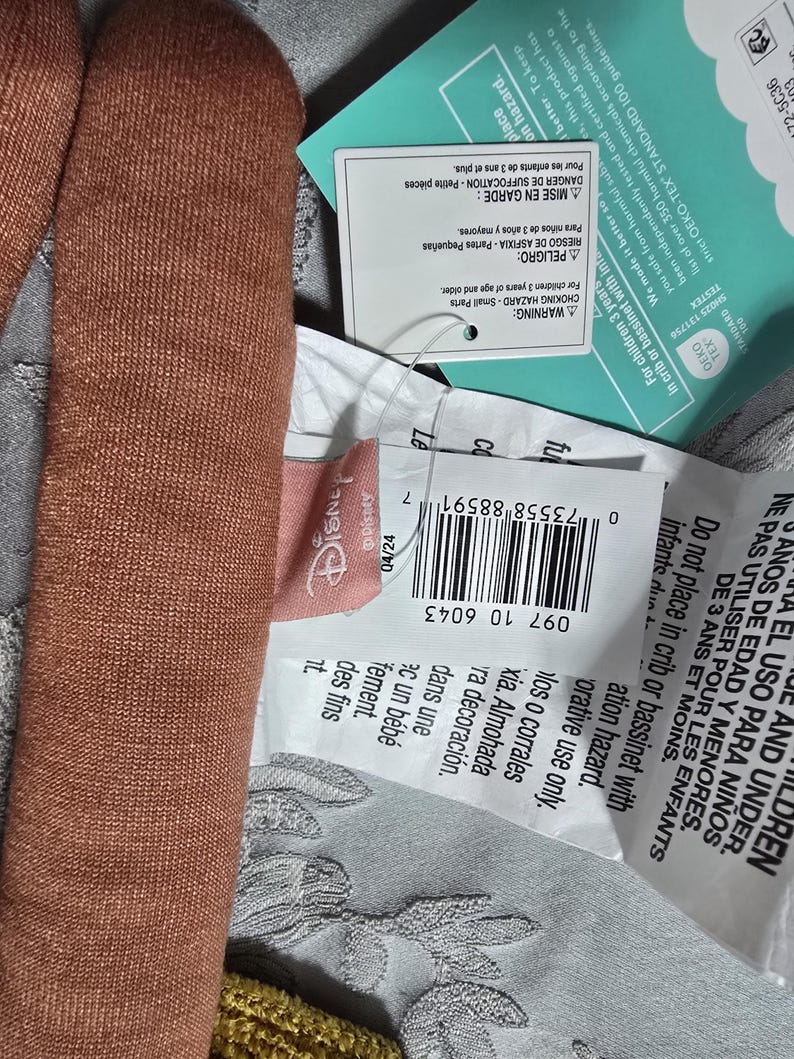 May include: Close-up of a brown plush toy with attached tags. The tags include the Disney logo, a barcode, and warnings in multiple languages about choking hazards for children.