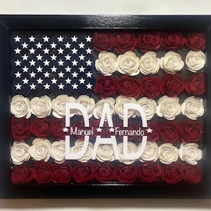 May include: A framed American flag made of red, white, and blue paper roses. The word "DAD" is written in white letters with the names "Manuel" and "Fernando" written below.