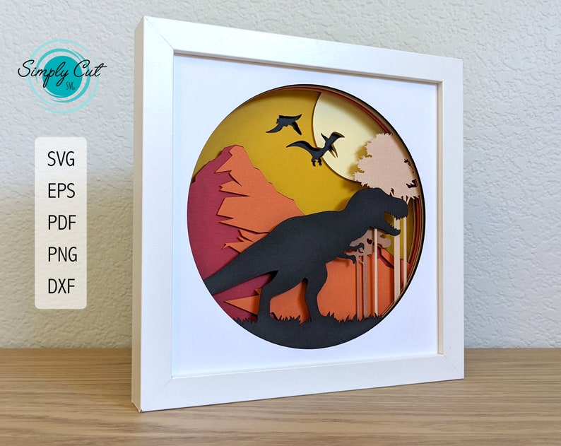 May include: A white frame containing a paper cut-out design of a T-Rex dinosaur in a sunset landscape. The dinosaur is black, the sunset is orange and yellow, and there are trees in the background.