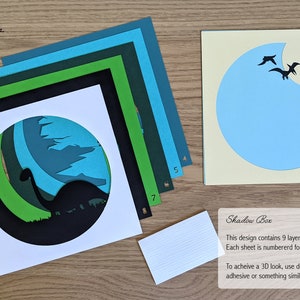 May include: A 9-layer papercraft shadow box kit featuring a dinosaur silhouette with a green, blue, and black colour scheme. The kit includes instructions for assembly and a note about using double-sided foam adhesive for a 3D effect.