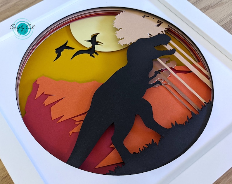 May include: A papercraft scene depicting a black silhouette of a Tyrannosaurus Rex standing in a forest with a yellow moon and two flying pterosaurs in the background. The scene is layered with different coloured paper, creating a 3D effect.