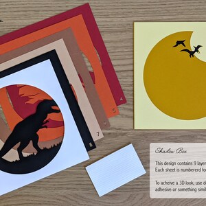 May include: A 9-layer papercraft shadow box kit featuring a silhouette of a dinosaur in a sunset scene. The kit includes 9 numbered sheets of paper, a white base, and instructions for assembly. To achieve a 3D look, use double-sided foam adhesive between each layer.
