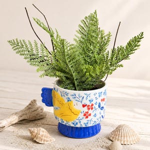 May include: A ceramic mug-shaped planter with a blue base and a yellow bird-shaped handle. The white exterior features a blue floral pattern with small red flowers. A green fern plant is potted inside the mug, which is surrounded by seashells.