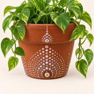 May include: A terracotta plant pot featuring a hand-painted mandala design made of white, pink, and purple dots. A trailing pothos plant with variegated green and yellow heart-shaped leaves grows out of the top of the round, tapered clay container.