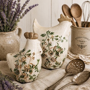 May include: Two cream-coloured ceramic chicken figurines decorated with a brown and green floral pattern. The chickens feature textured brown combs and sit on a light-coloured fabric surface alongside two metal spoons and a ceramic utensil crock.