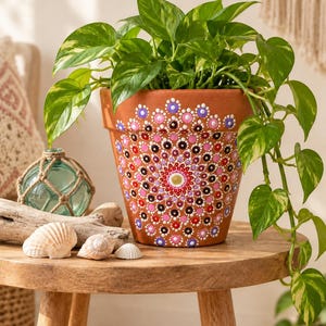 May include: A terracotta plant pot featuring a hand-painted mandala design with concentric circles of dots in shades of pink, purple, white, and black. A vibrant green pothos plant with variegated leaves grows from the pot, which sits on a wooden table.