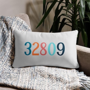 May include: White rectangular pillow with the number "32809" printed in teal, orange, light blue, and navy blue. The pillow is resting on a patterned blanket and a gray armchair.