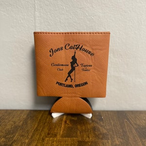 May include: Brown leather can cooler with the text "Jons Cat House" in black lettering. The text "Gentleman's Club" and "Topless Tastee" are also printed in black. A silhouette of a woman in a suggestive pose is printed on the cooler. The text "Portland, Oregon" is printed at the bottom.