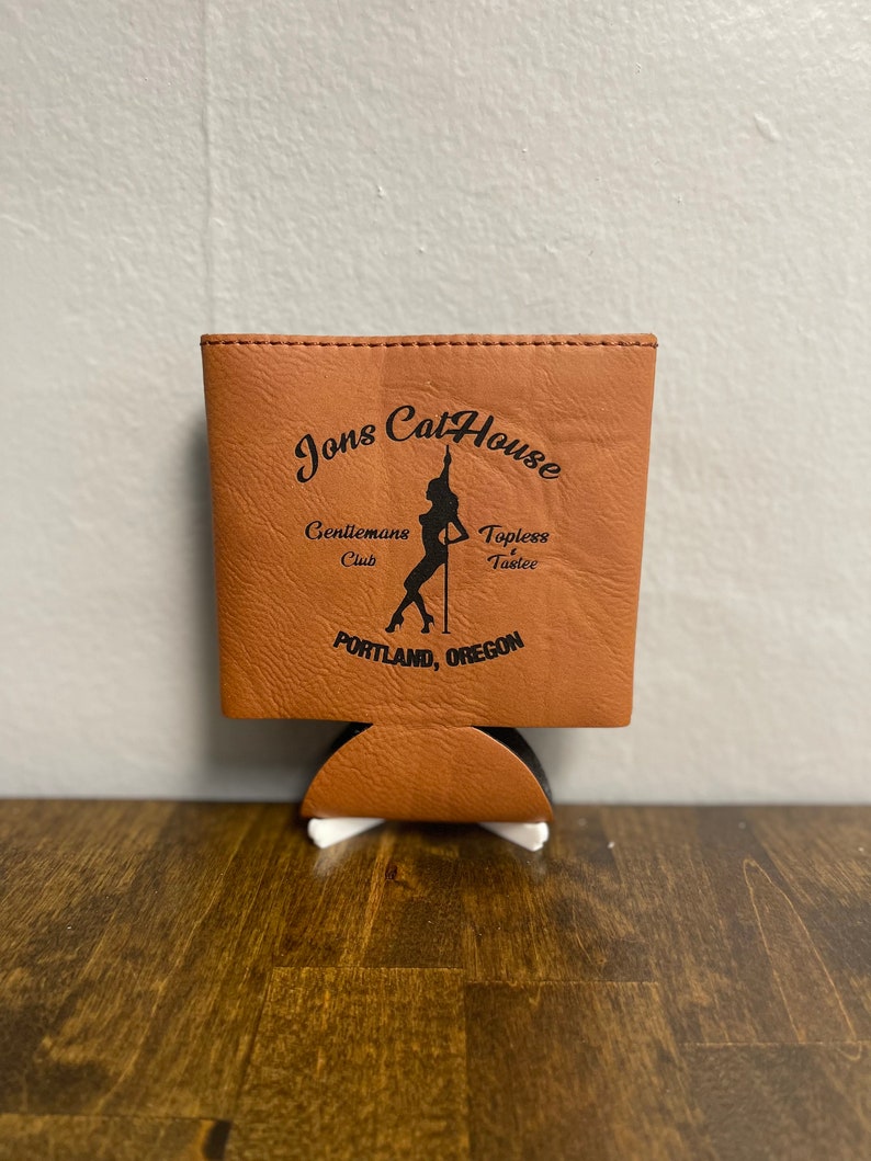May include: Brown leather can cooler with the text "Jons Cat House" in black lettering. The text "Gentleman's Club" and "Topless Tastee" are also printed in black. A silhouette of a woman in a suggestive pose is printed on the cooler. The text "Portland, Oregon" is printed at the bottom.
