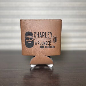 May include: Brown leather can cooler with a black and white graphic of a man with a beard and sunglasses wearing a baseball cap. The graphic says "Charley the Plumber" and includes a YouTube logo.