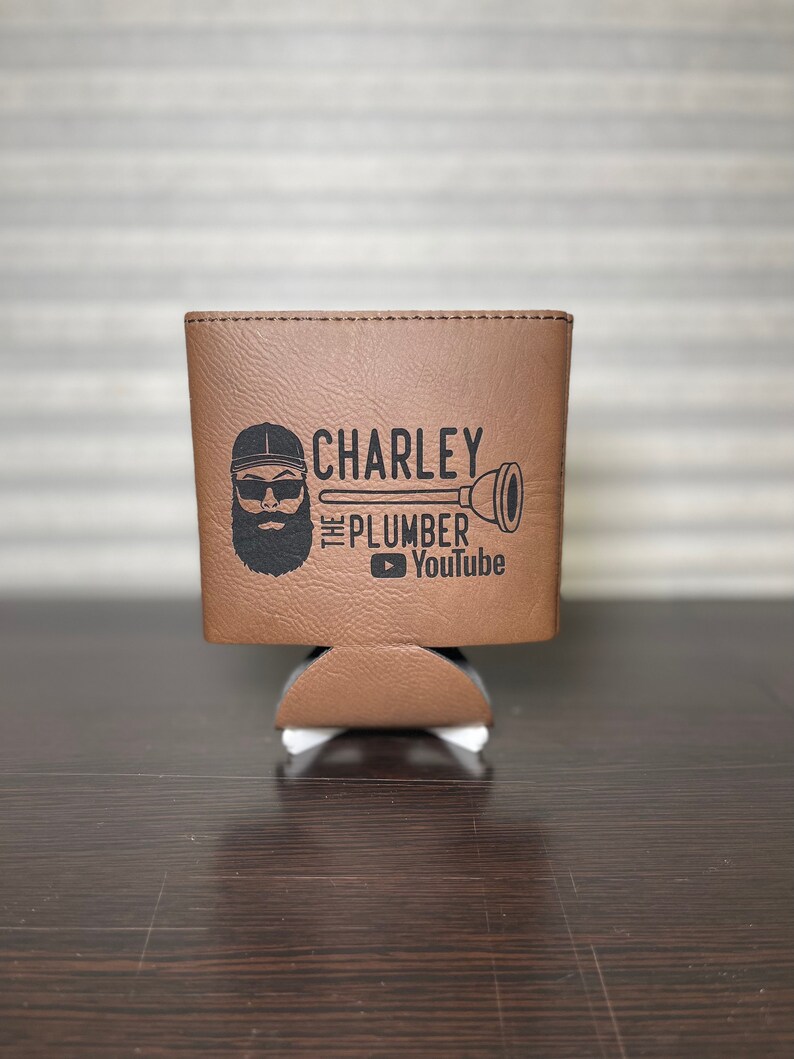 May include: Brown leather can cooler with a black and white graphic of a man with a beard and sunglasses wearing a baseball cap. The graphic says "Charley the Plumber" and includes a YouTube logo.