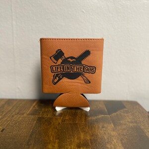 May include: A brown leather can cooler with a black graphic of crossed axes and a chainsaw with the text "LIFE IN THE 906".