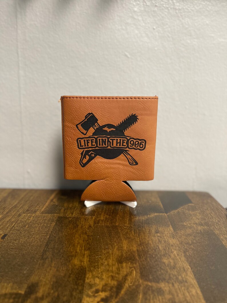 May include: A brown leather can cooler with a black graphic of crossed axes and a chainsaw with the text "LIFE IN THE 906".