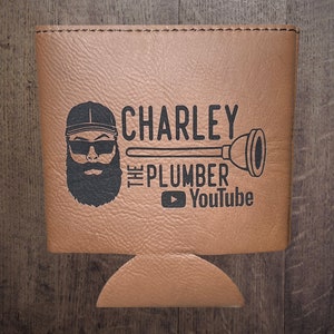 May include: Brown leather can cooler with a black and white graphic of a man with a beard wearing a baseball cap and sunglasses. The graphic also includes the text "CHARLEY THE PLUMBER" and a YouTube logo.