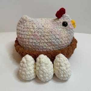 May include: A crocheted plush chicken with a red comb and yellow beak sitting in a brown crocheted nest with three white crocheted eggs in front of it.