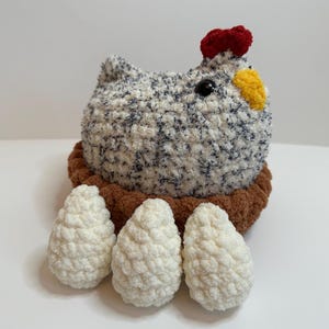 May include: A handmade crochet chicken with a red comb, yellow beak, and black eye, sitting in a brown nest. Three white crochet eggs are in front of the chicken. The chicken is white with blue and black flecks.