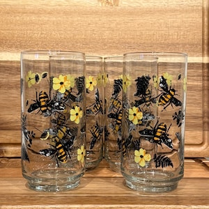 May include: Set of four clear drinking glasses with a bee and floral design. The glasses feature black and yellow bees, yellow flowers, and green fern-like leaves. The glasses are displayed on a wooden surface.