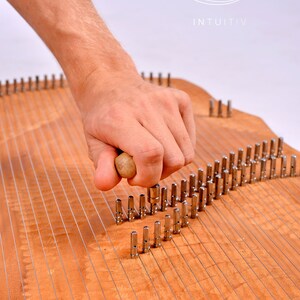 May include: A wooden musical instrument with metal pins and strings. A hand is holding a wooden mallet and striking the pins.