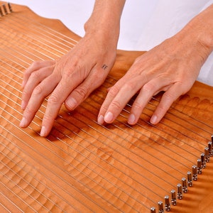 May include: A person's hands playing a wooden lyre with strings. The lyre has a carved wooden surface and metal tuning pegs.