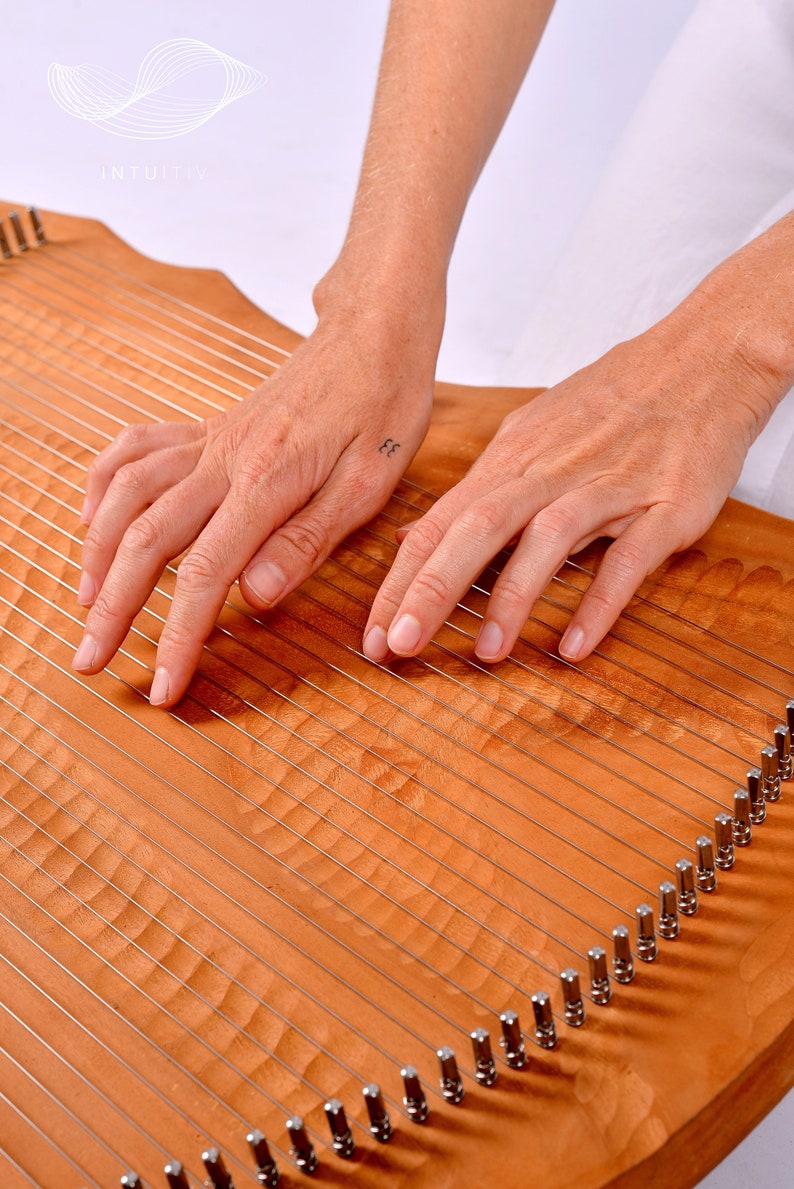 May include: A person's hands playing a wooden lyre with strings. The lyre has a carved wooden surface and metal tuning pegs.