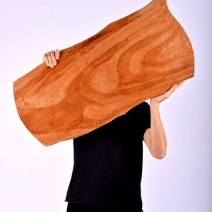 May include: A person holds a large, brown, wooden board with a unique grain pattern. The board is held in front of the person's face, obscuring their features. The background is a plain white.