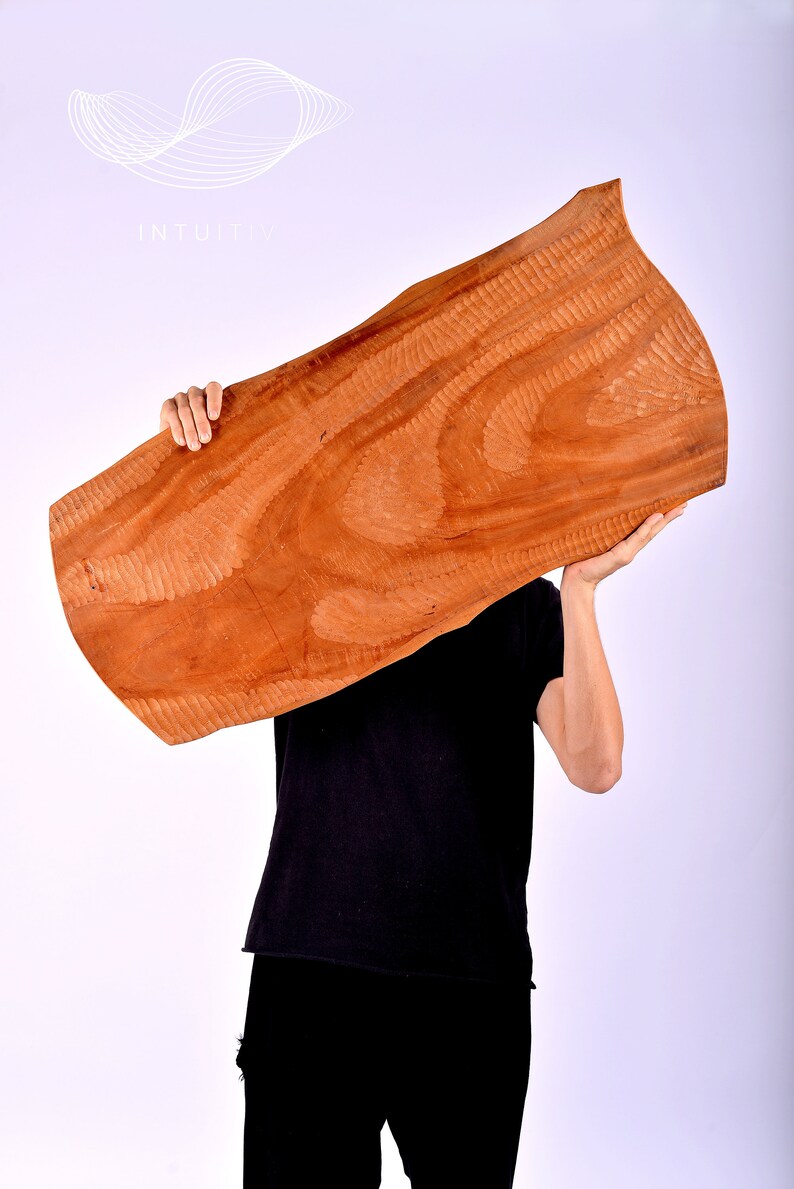 May include: A person holds a large, brown, wooden board with a unique grain pattern. The board is held in front of the person's face, obscuring their features. The background is a plain white.