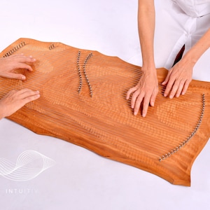 May include: Two people are playing a wooden sound healing instrument called a  'Lyre' or 'Sound Board'. The instrument has a natural wood finish and is covered in metal pins. The instrument is resting on a white surface. The text 'INTUITIV' is visible in the lower left corner of the image.