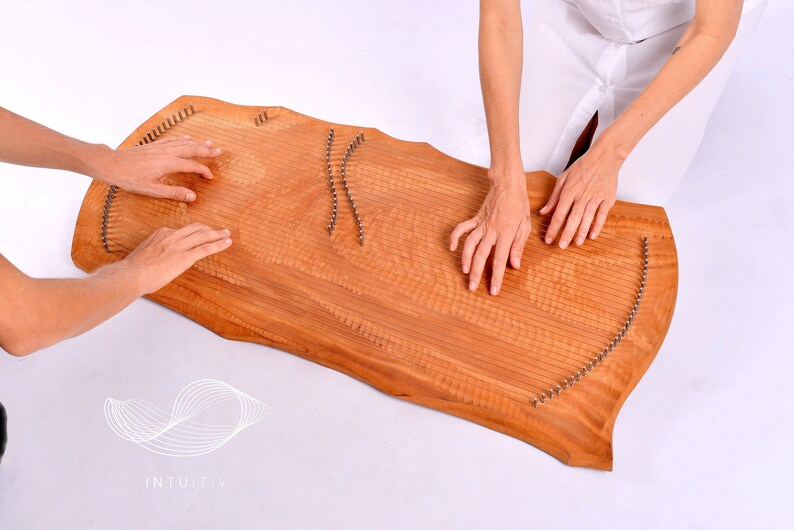 May include: Two people are playing a wooden sound healing instrument called a  'Lyre' or 'Sound Board'. The instrument has a natural wood finish and is covered in metal pins. The instrument is resting on a white surface. The text 'INTUITIV' is visible in the lower left corner of the image.