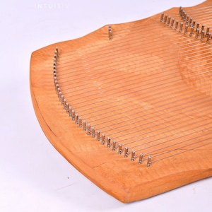 May include: A wooden lyre with a curved shape and multiple strings attached to metal tuning pegs. The lyre is made of light wood with a natural finish.