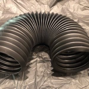 Op de afbeelding: Zwarte flexibele gegolfde buis, gemaakt van plastic, met een diameter van ongeveer 10 cm. De buis is gebogen in een U-vorm.