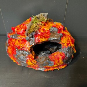 May include: A small, ceramic cave painted with red, orange, and gray paint. The cave has a small opening and a green moss-like material on top. The text "MHHLM2" is printed on a white label below the cave.