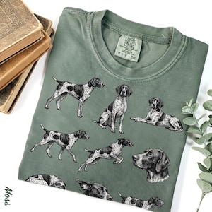 May include: Sage green t-shirt featuring multiple black and white illustrations of a dog breed. The illustrations depict the dogs in various poses, including standing, sitting, and lying down. The shirt has a crew neck and a tag that reads "Comfort Colors".