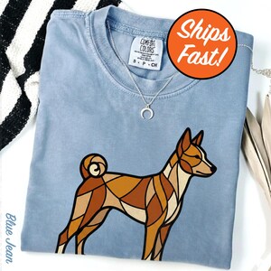 May include: A blue jean colored t-shirt featuring a geometric, stained-glass style illustration of a brown and tan dog with a curled tail. The shirt has a crew neckline and a white tag at the collar. A silver crescent moon necklace rests on the fabric.